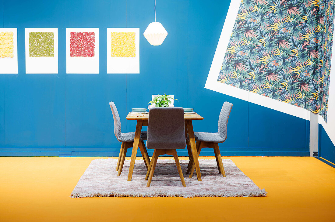 Ein Tisch und sechs Stühle stehen auf einem Teppich. Der Boden ist gelb, die Wand ist blau.